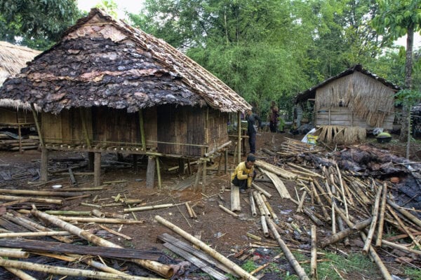 thailand hilltribe karen man kneeling next to a grass-roofed bamboo house